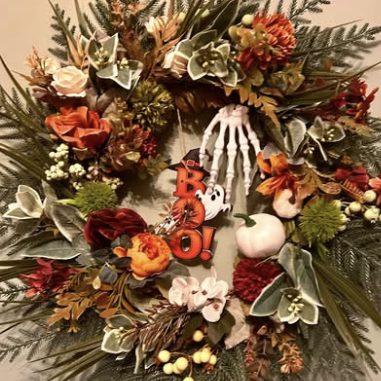 Halloween wreath of red and orange flowers and pine, with a skeleton hand and BOO! lettering