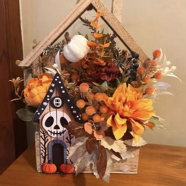 Wooden house-shaped box of orange flowers and pumpkins, with a small skull-faced house at the front